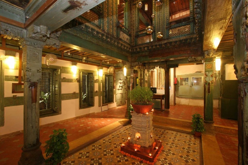 Ornately carved teak balconies and pillars encircle a lantern-lit courtyard lobby with patterned tile flooring and a potted plant centrepiece.