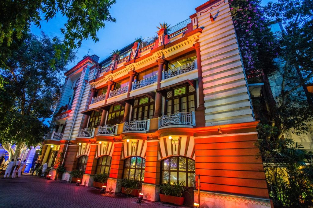 At dusk, the House of MG's terracotta and cream three-storey facade glows beneath uplighting, its balconies and columned windows framed by trees.