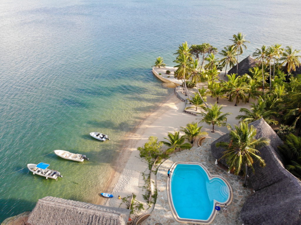 Aerial view of Manda Bay Lodge's palm-lined beach, swimming pool, thatched roofs and three motorboats moored in clear shallows.
