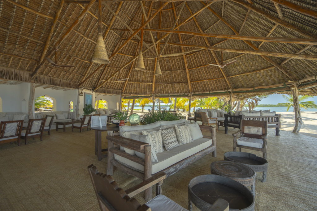 Open-sided lounge and bar under a high makuti thatched roof, with cushioned sofas and carved wooden tables, and palms and sea beyond.