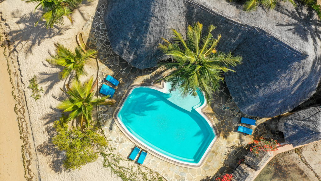 Overhead view of the swimming pool with blue loungers, a flagstone deck, coconut palms and thatched roofs at Manda Bay Lodge, Kenya.