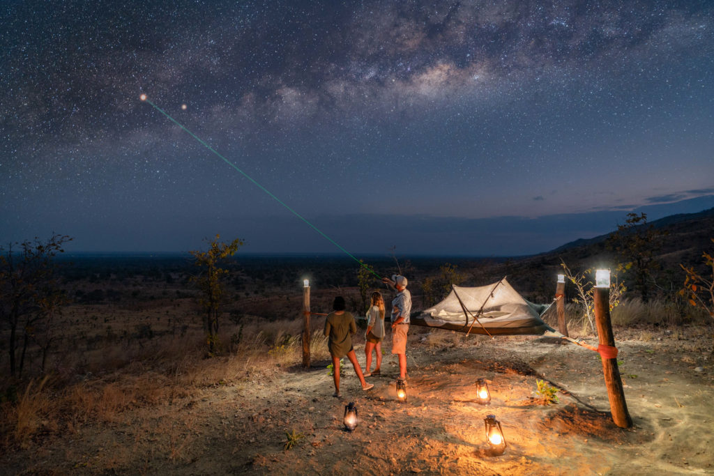 Guests stargaze beside a starbed under the Milky Way, lit by lanterns on a hilltop.