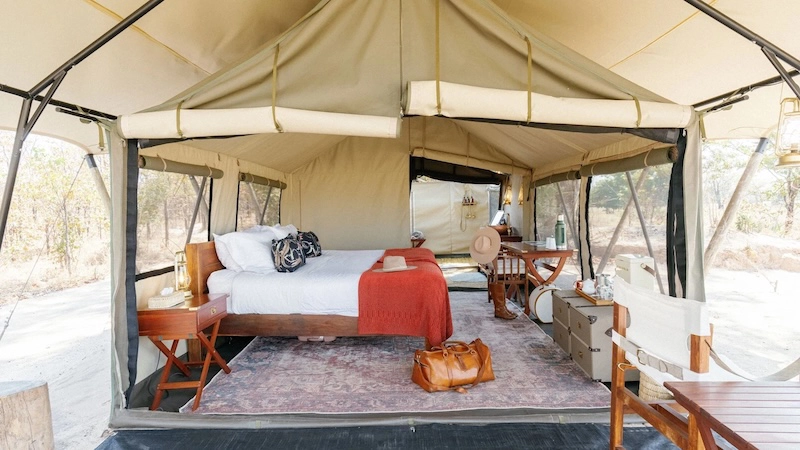 The interior of a classic safari tent at Time + Tide Luwi Camp in South Luangwa National Park, featuring a king bed, timber furnishings and views out to the surrounding woodland.