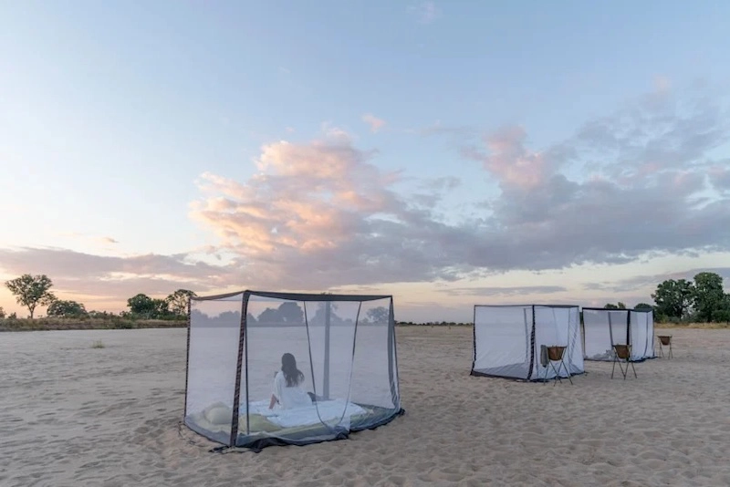 A private sleep-out experience at Time + Tide Luwi Camp in South Luangwa National Park, with mesh beds arranged on the riverbank beneath a pastel sunset sky.