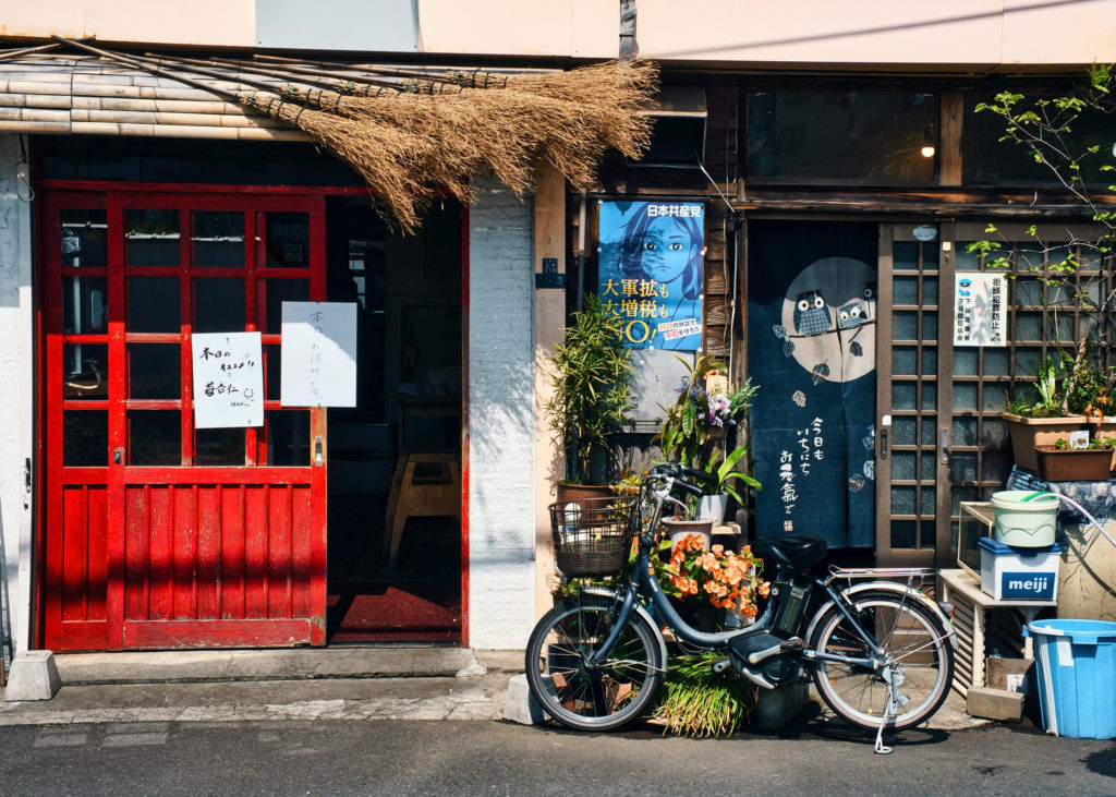 A red wooden door and a bicycle sit beside potted flowers and a navy noren curtain decorated with owls, outside an old shopfront in a Tokyo backstreet.