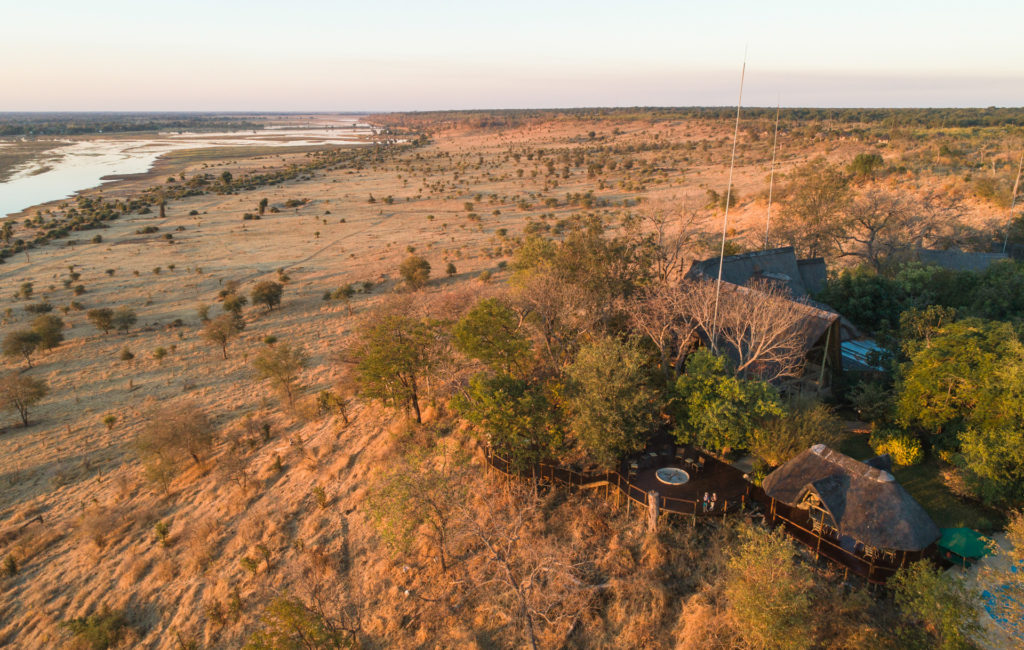 An aerial view of Muchenje Safari Lodge perched on the escarpment above the Chobe River, its thatched main area and circular viewing deck surrounded by woodland and open floodplains.
