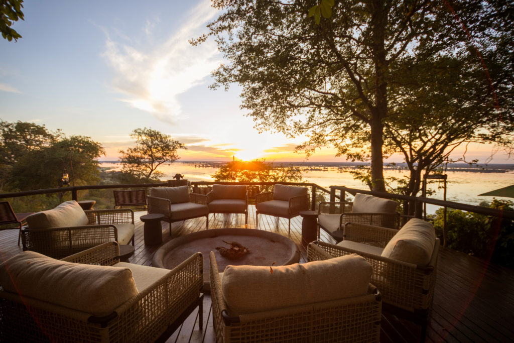 The main viewing deck at Muchenje Safari Lodge at sunset, with cushioned rattan chairs arranged around a central fire bowl and the Chobe River burning gold on the horizon.