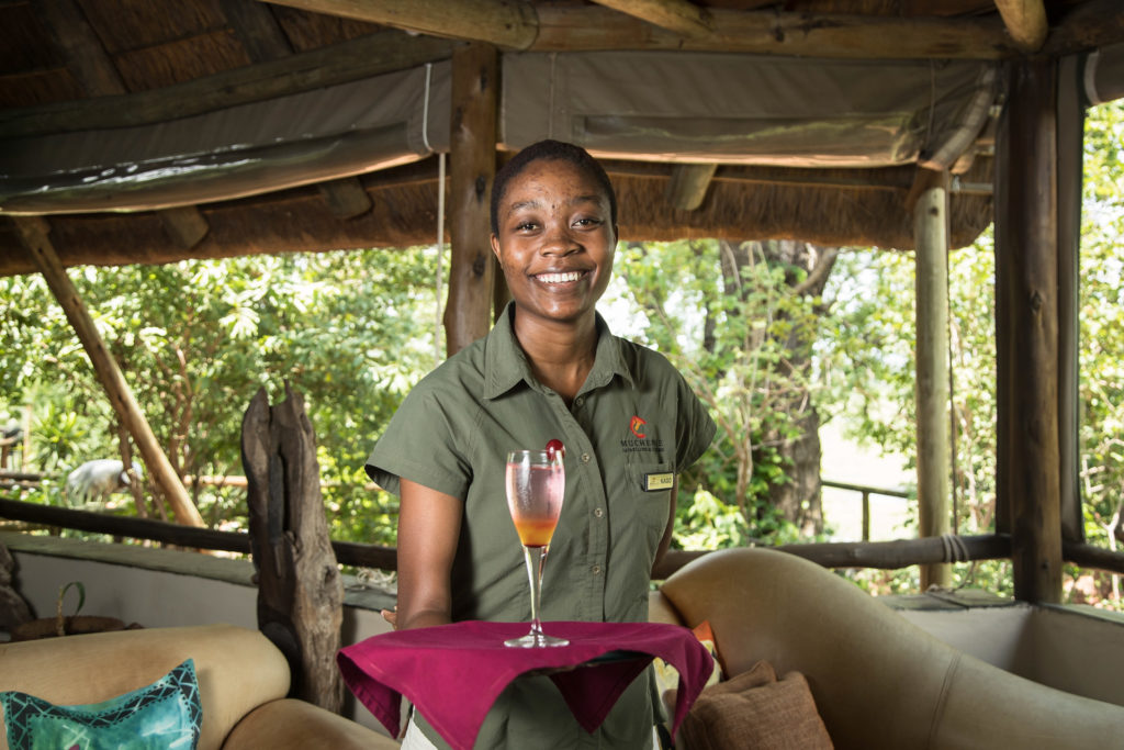 A smiling Muchenje staff member holds a cocktail on a tray in the open-air lodge bar, with the green canopy of the Chobe Forest Reserve visible behind her.