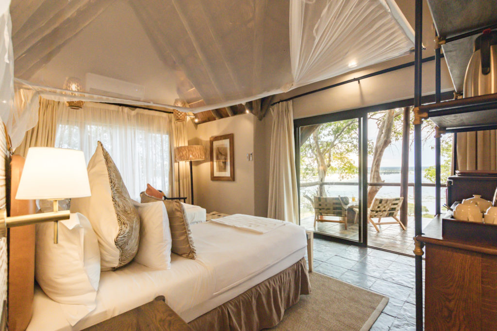 A canopied king bed in a Muchenje Safari Lodge cottage, with warm gold tones, wildlife artwork and sliding glass doors opening to a private deck overlooking the Chobe River.