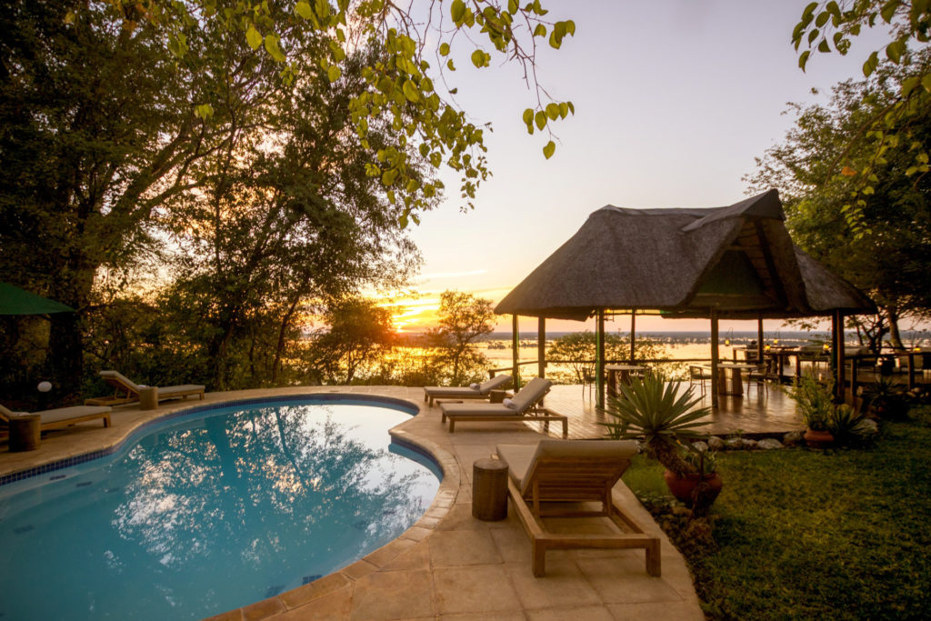 The Muchenje Safari Lodge swimming pool at sunset, with teak sunloungers along the pool's edge, a thatched bar pavilion and the Chobe River glowing amber in the background.