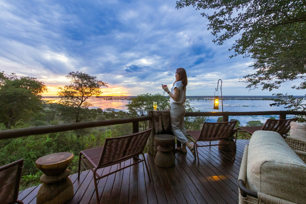 A guest stands on Muchenje Safari Lodge's timber viewing deck at sunset, a paraffin lantern glowing beside her as the Chobe River catches the last light of the day.