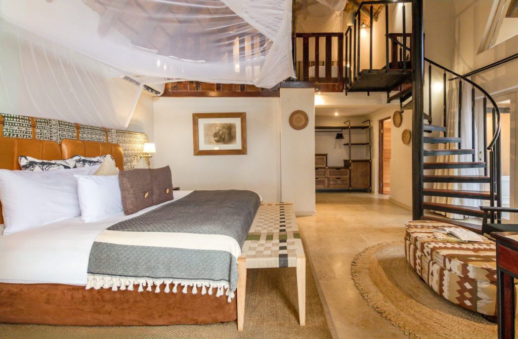 The family cottage interior at Muchenje Safari Lodge, featuring a canopied king bed with a leather and batik headboard, woven bench, kilim-print ottoman and a spiral staircase rising to a mezzanine sleeping loft.