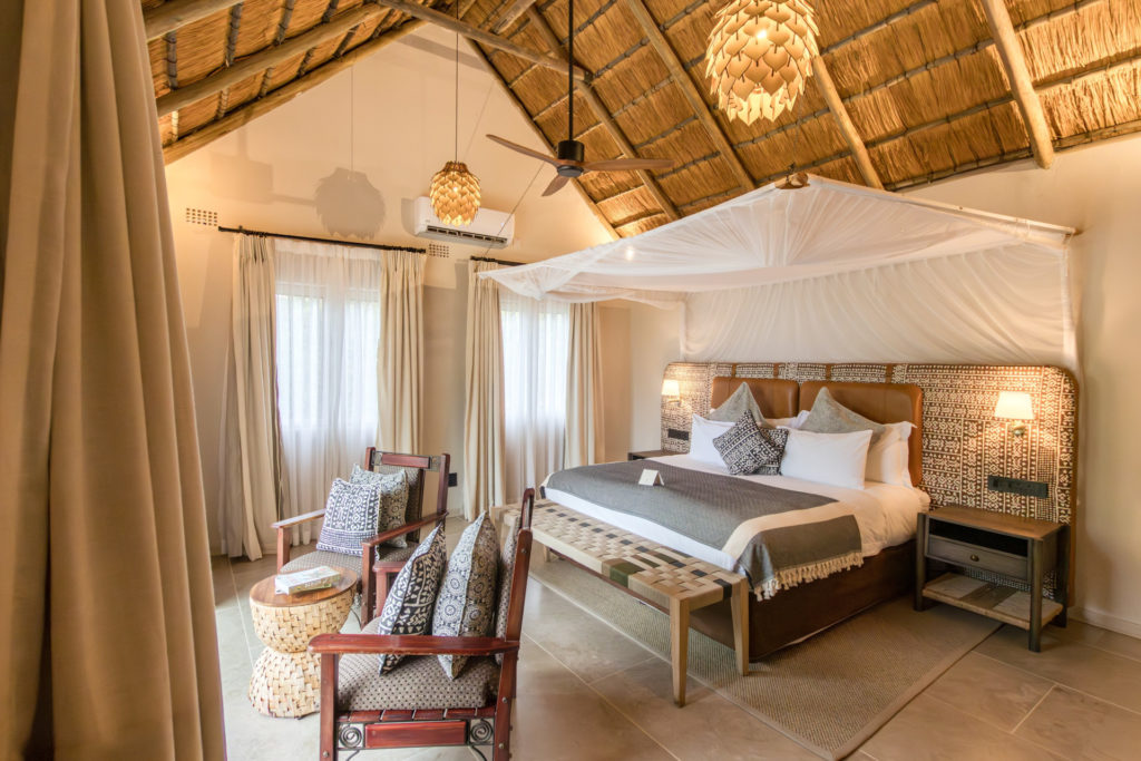 A spacious Muchenje cottage interior with a canopied king bed, leather and batik headboard, artichoke pendant lights, thatched ceiling and a seating area beside sheer-curtained windows.