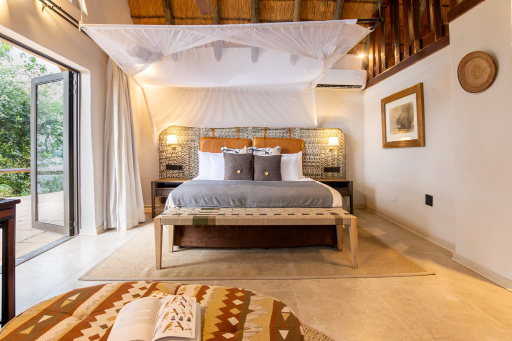 A canopied king bed in a Muchenje Safari Lodge cottage with a leather and batik headboard, woven bench and open bifold doors leading to a private deck among the trees.