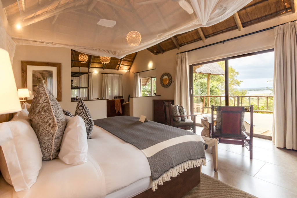 A spacious Muchenje cottage bedroom with a canopied king bed, patterned cushions, thatched ceiling and sliding doors opening to a private balcony with Chobe River views.
