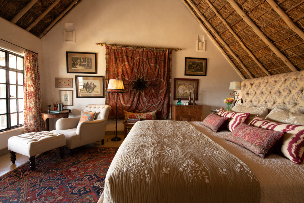 West Wing bedroom with an upholstered headboard, embroidered bedspread, paisley and ikat cushions and a thatched roof, Olepangi Farm, Kenya.