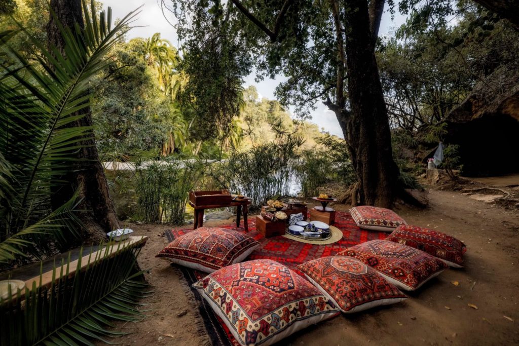 Picnic lunch set on red kilim rugs and embroidered floor cushions beside a palm-fringed pool, with dishes laid out on low wooden boxes.