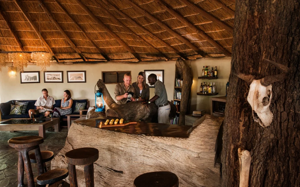 Guests and a guide review wildlife photographs at the rustic driftwood bar inside the thatched main lodge at Kaingo Camp, another couple relaxing on sofas in the background, South Luangwa, Zambia.