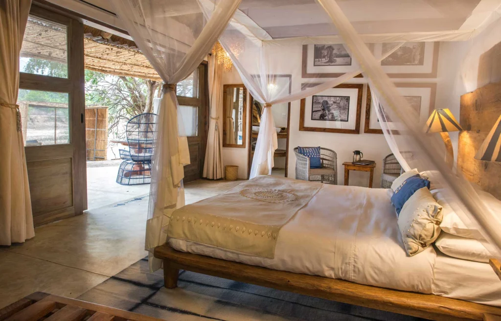A light-filled Kaingo Camp chalet interior featuring a canopied four-poster bed with crisp white linen, shell-embroidered throw, geometric wall art, black-and-white wildlife prints in timber frames, and open doors leading to a private deck with wicker seating.