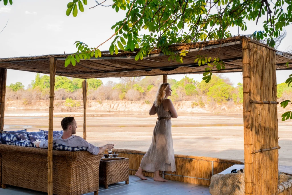 A couple relax on a reed-and-bamboo riverside deck at Kaingo Camp, the woman gazing out over the wide sandy Luangwa River channel and green treeline beyond in warm afternoon light, Zambia.