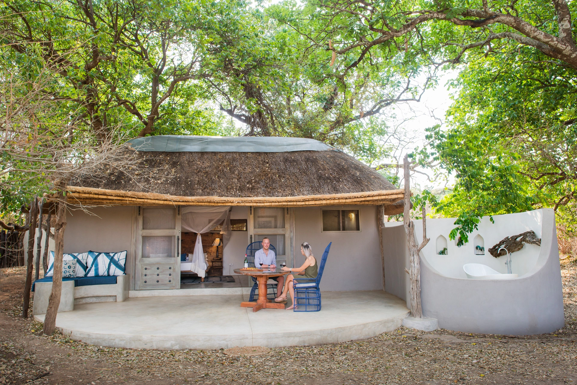 A couple share wine at a round table on the private veranda of a thatched chalet at Kaingo Camp, the open bedroom with canopied bed visible behind them and an outdoor bathtub tucked to one side, South Luangwa, Zambia.