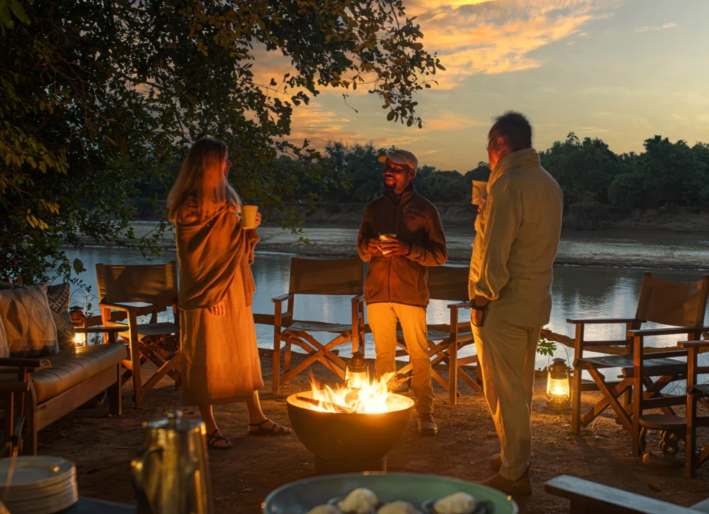 Guests and a guide warm their hands around a glowing fire bowl on the riverbank at Kaingo Camp, lanterns flickering beside safari chairs as the Luangwa River reflects a golden sunset sky behind them, Zambia.