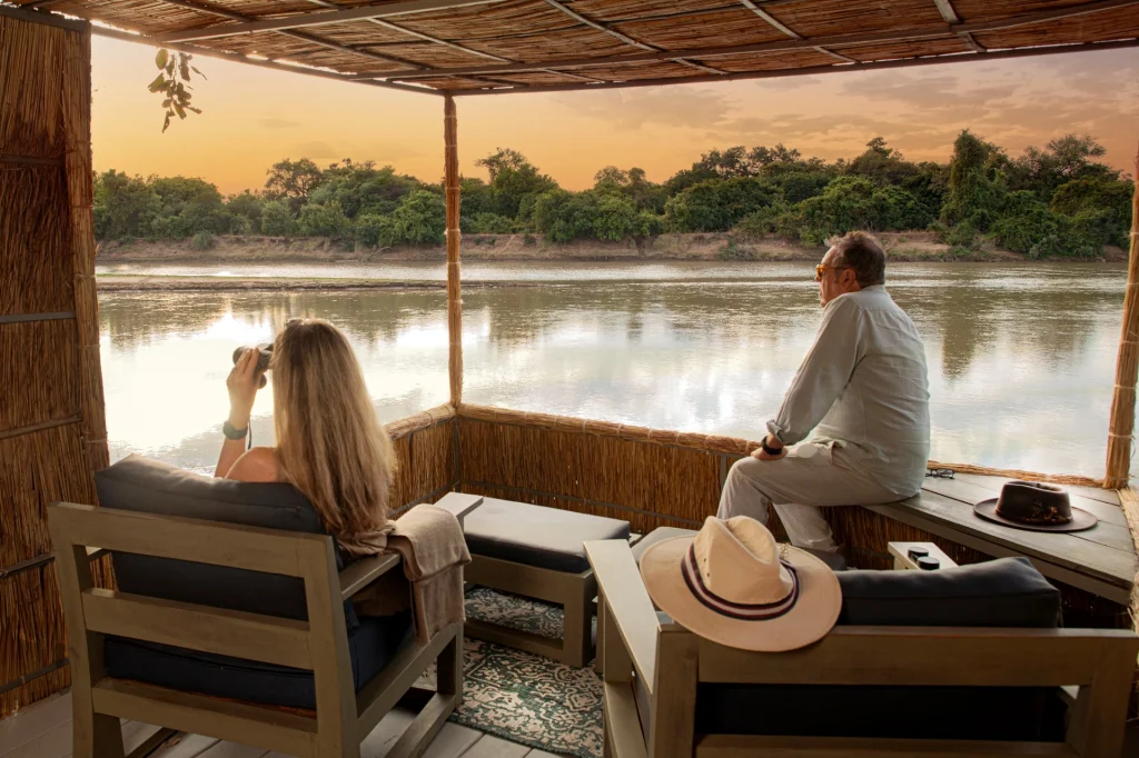 Two guests relax on a reed-screened riverside viewing platform at Kaingo Camp, one scanning through binoculars as the Luangwa River glows amber in the fading evening light, South Luangwa, Zambia.