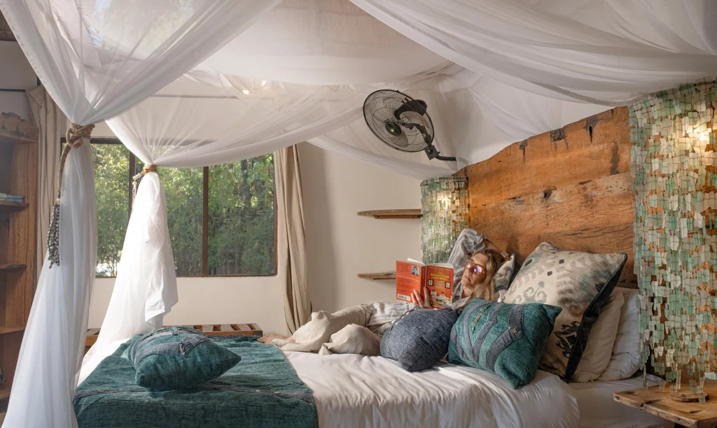 A woman reclines on a four-poster bed draped with white mosquito netting in a Kaingo Camp chalet, reading a book amid teal and grey cushions, with a rustic wooden headboard and mosaic-tiled wall feature visible alongside bush views through large windows.