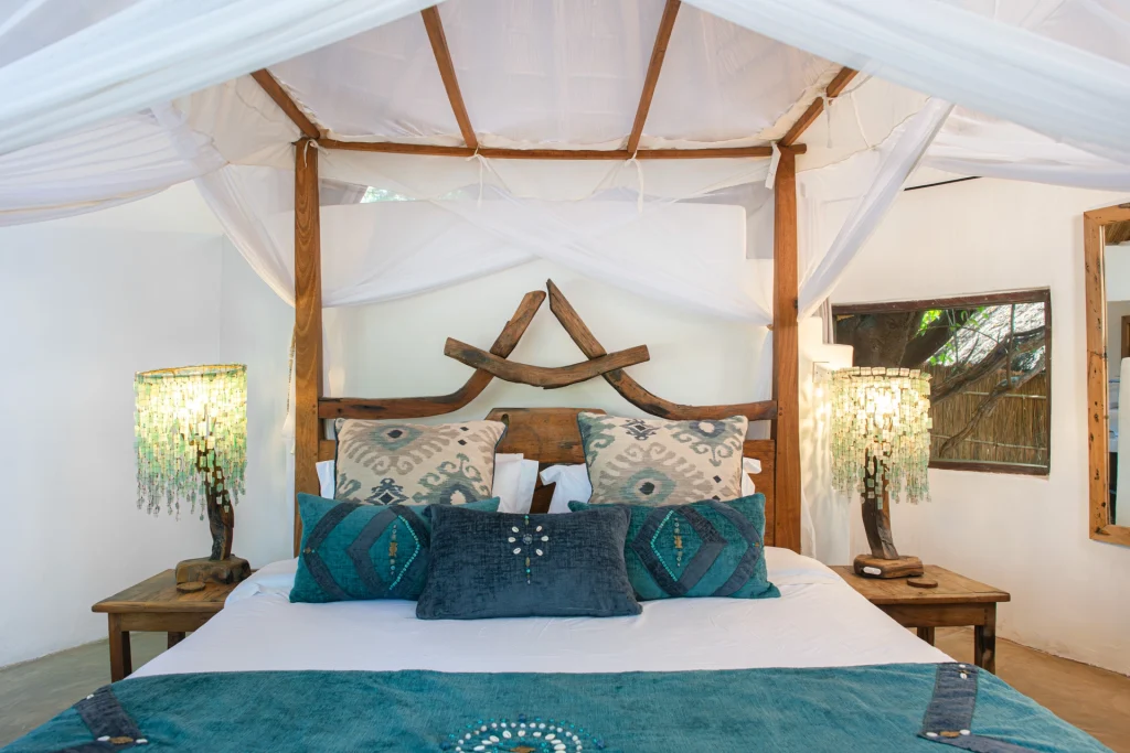 A beautifully dressed four-poster king bed in a Kaingo Camp chalet, featuring a sculptural driftwood headboard, teal velvet and ikat cushions, and two glowing capiz shell bedside lamps, framed by white mosquito netting and timber canopy posts.
