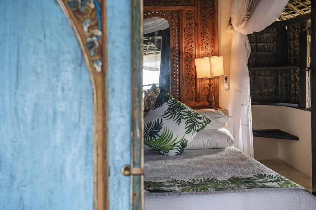 View through a weathered blue carved door to a bed with palm-print cushions, a carved headboard and a bedside lamp.