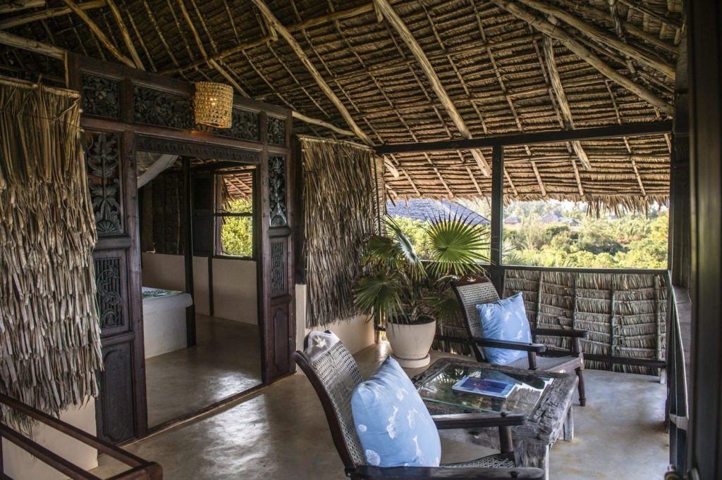 Thatched upstairs veranda with cane chairs, blue cushions and a potted palm, opening through carved doors into a bedroom.