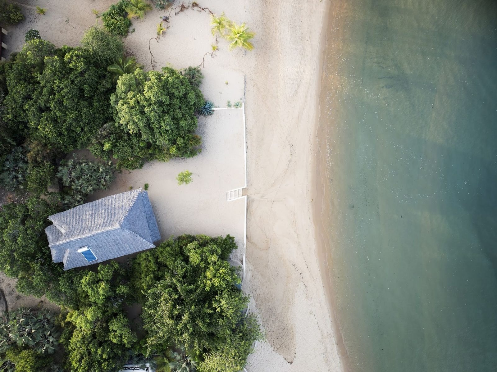 Aerial view of a thatched beach house among coastal trees above white sand and shallow green water on Lamu, Kenya.