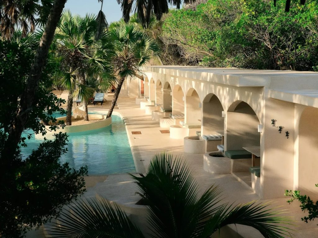 Whitewashed arched alcoves with cushioned seating line a palm-shaded pool at The Cabanas Lamu in dappled light.