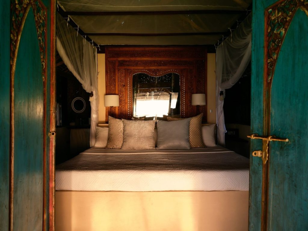 Turquoise carved doors open onto a four-poster bed with silk cushions, mosquito net and a mirrored headboard in soft light.