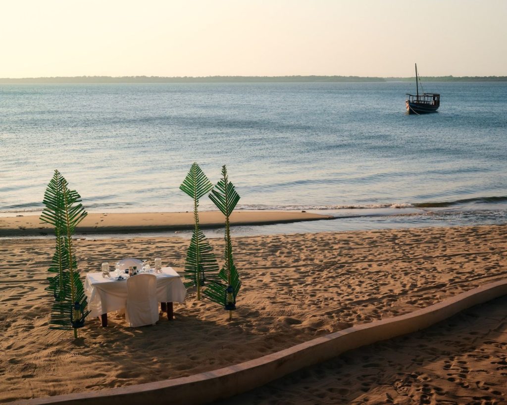 Private beach dinner table set between palm frond screens and lanterns, with a dhow anchored offshore at dusk in Lamu.