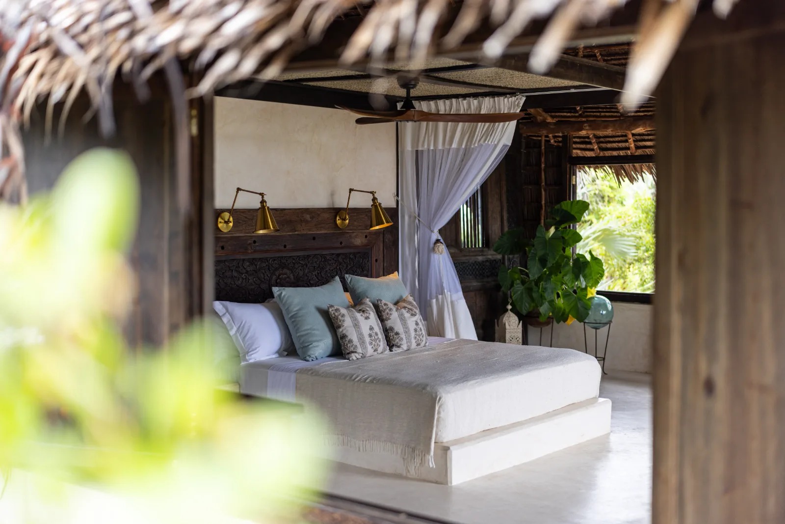Bedroom with a raised plaster bed, brass wall lamps, carved headboard and ceiling fan, with greenery outside the window.