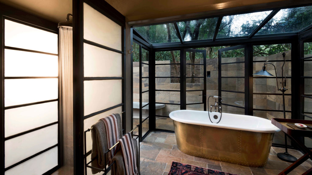 A freestanding copper bathtub sits beneath a glass roof beside an open-air outdoor shower