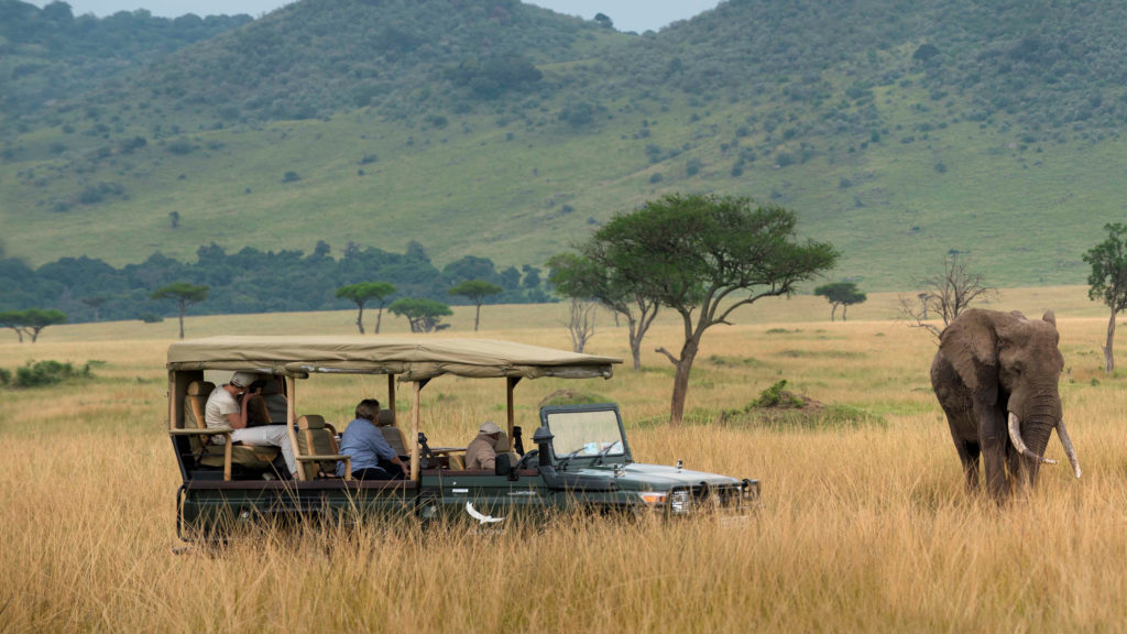 A safari vehicle stops near a grazing elephant against a backdrop of forested hills