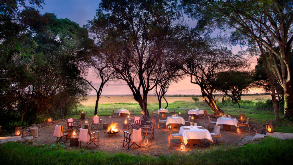 Tables set with white linen and lantern light gather beneath acacia trees for a bush dinner at Bateleur Camp