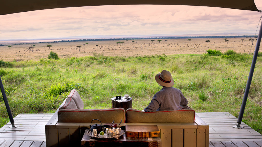 A guest looks out from a private veranda daybed over plains dotted with grazing wildebeest