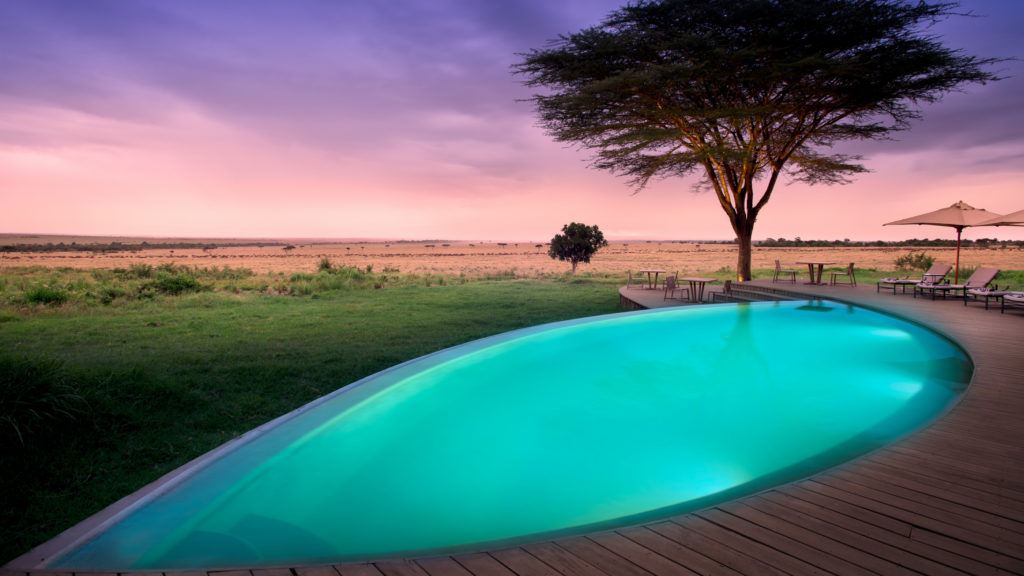 An infinity pool glows turquoise at dusk beside acacia trees and the open plains of the Maasai Mara