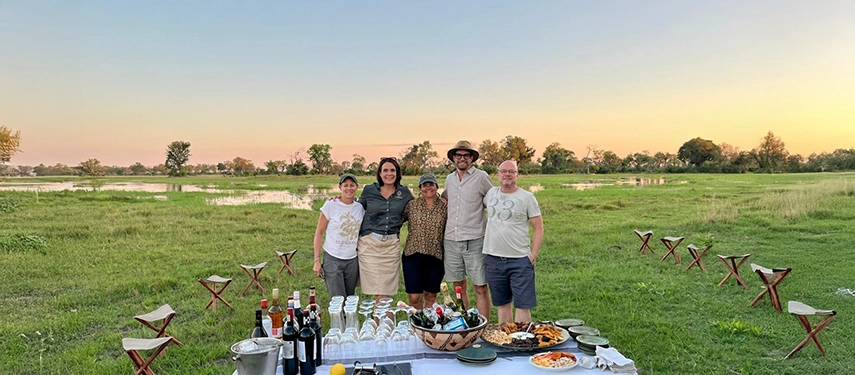 Safari guests and hosts pose beside a bush dining table set with drinks and canapés, celebrating a Karangoma sundowner in open floodplains at dusk.