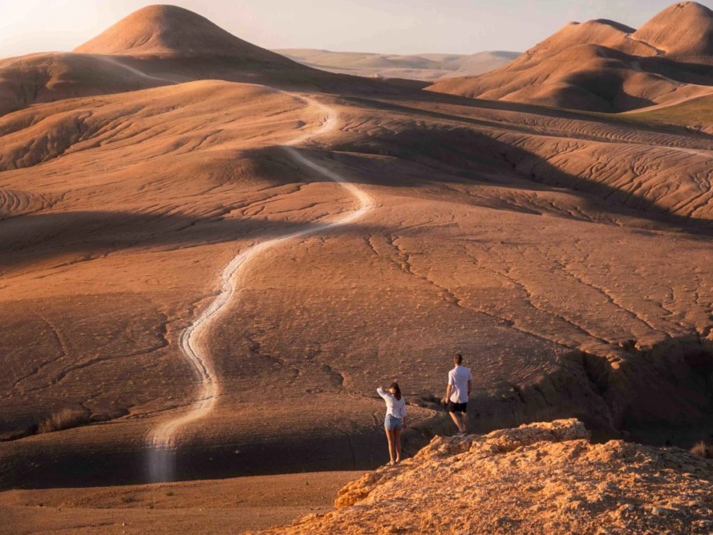 Two travellers pause on a ridge above the Agafay Desert, a pale trail winding through the ochre hills below.