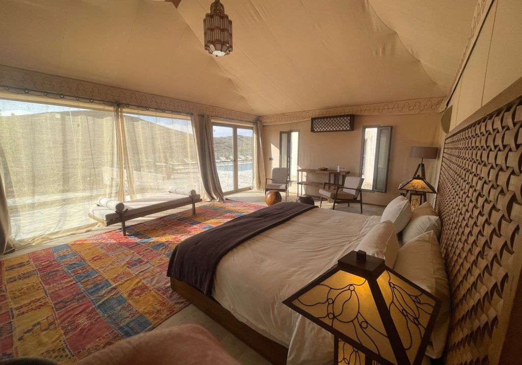 A lodge bedroom with a patchwork rug and sheer curtains opening onto a private pool and desert view.