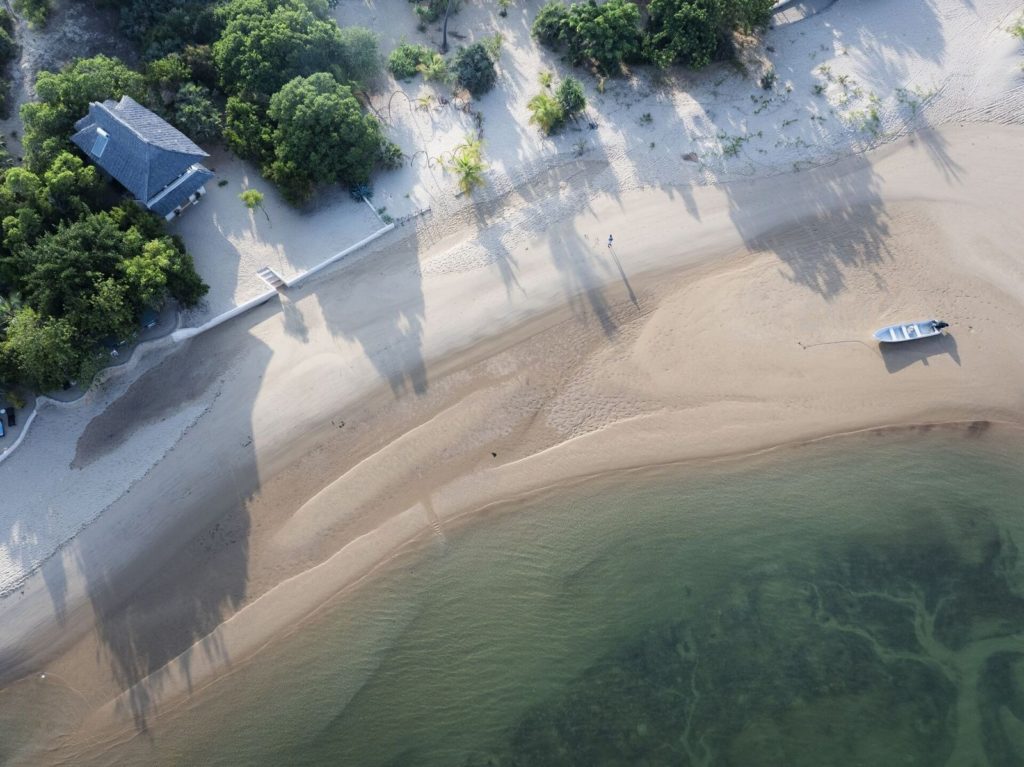 Aerial view of palm shadows stretching across a wide sandy beach, with a small boat pulled up below a thatched house.