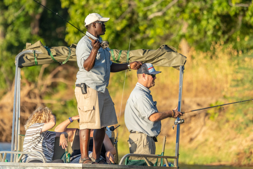 Guests fish from a boat on the Zambezi as their guide watches the line at sunset.