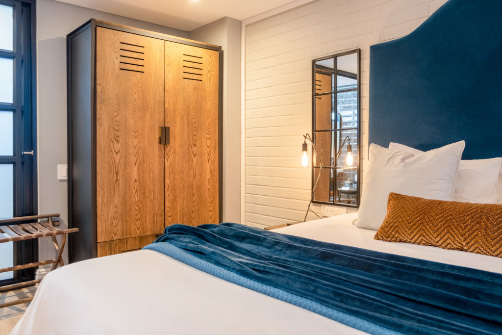 Bedroom at The Weinberg with timber wardrobe, blue upholstered headboard and exposed brick feature wall
