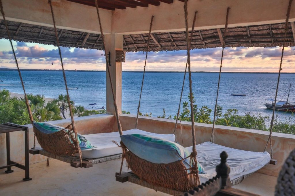 Rope-hung swing beds on a thatched rooftop terrace overlooking the Lamu channel, with dhows and kitesurfers at sunset.