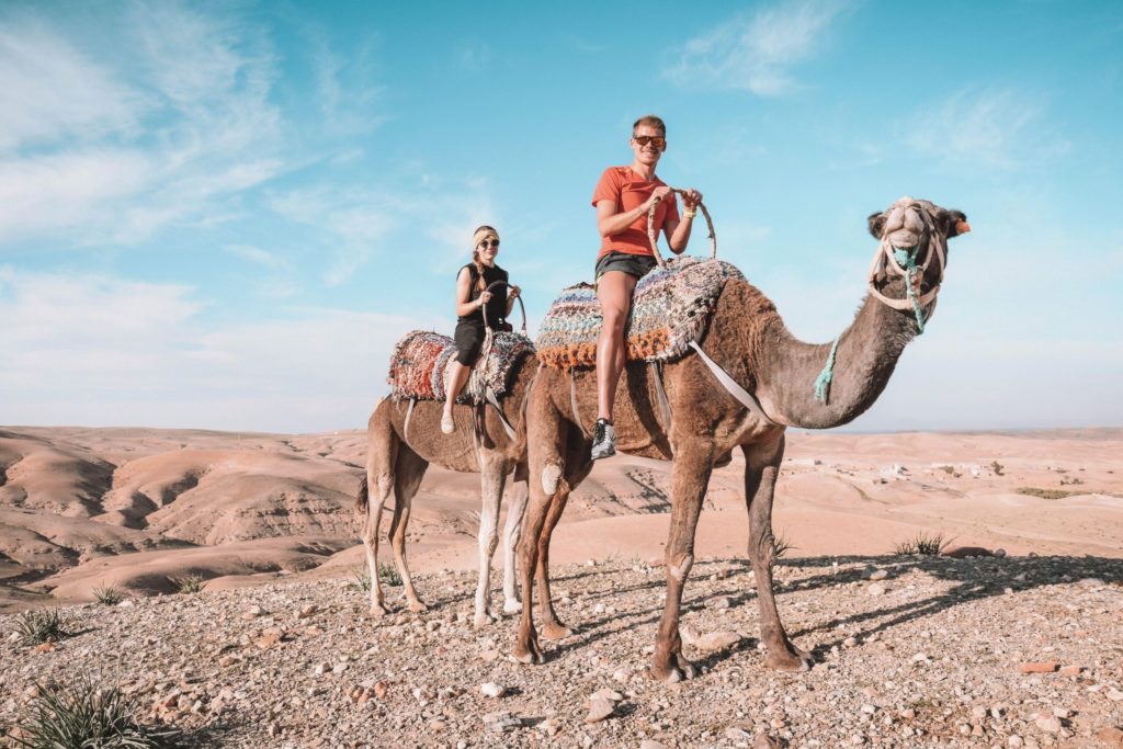 A couple ride camels across the folded hills of Agafay, the desert unrolling behind them.