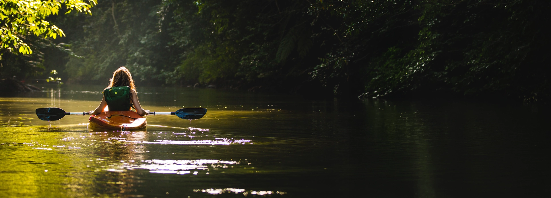 A woman kayaks alone along a calm jungle river, dappled golden light filtering through the dense forest canopy above and glittering on the still water ahead.